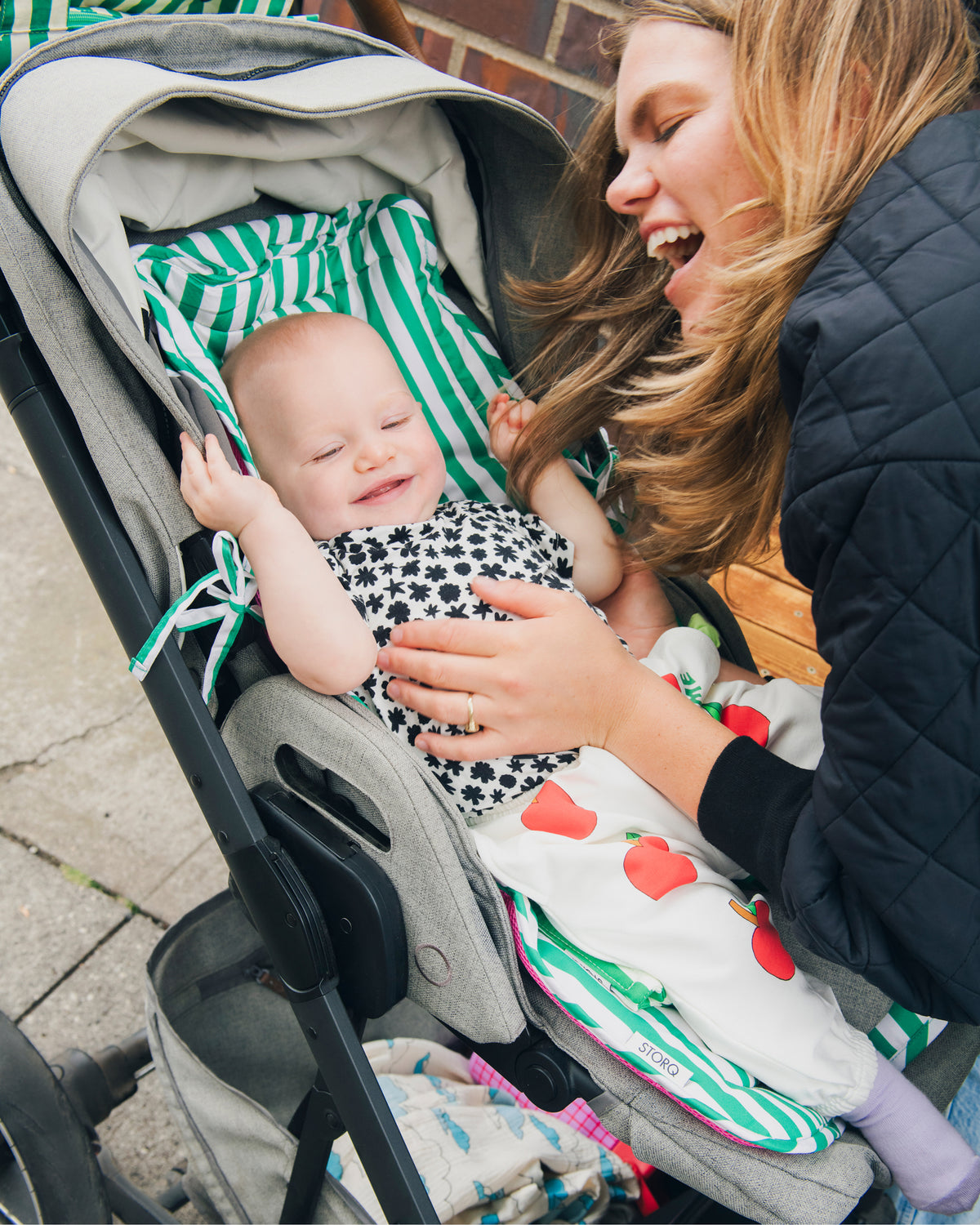 A baby sitting in a gray stroller on a Stroller Party™ Seat Liner & Strap Covers in Green Stripe, wearing Baby Onesie in Cream Chamomiles and French Terry Jogger in Ivory Apple, smiling as a woman in a black quilted jacket leans in close. The background is an outdoor setting on a city sidewalk.