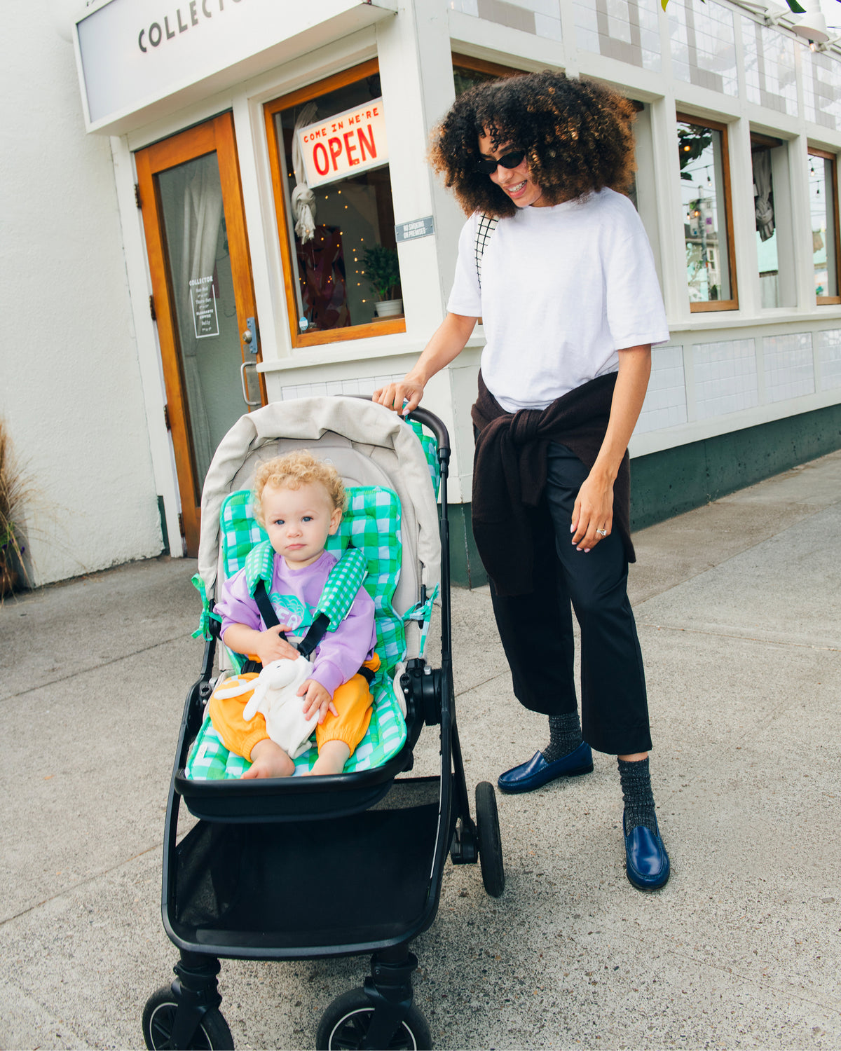 Model is pushing baby in a beige stroller on the Stroller Party™ Seat Liner & Strap Covers in Teal Gingham, while wearing French Terry Jogger in Orange Bird and Graphic Sweatshirt in Purple Bird, in a downtown sidewalk setting.