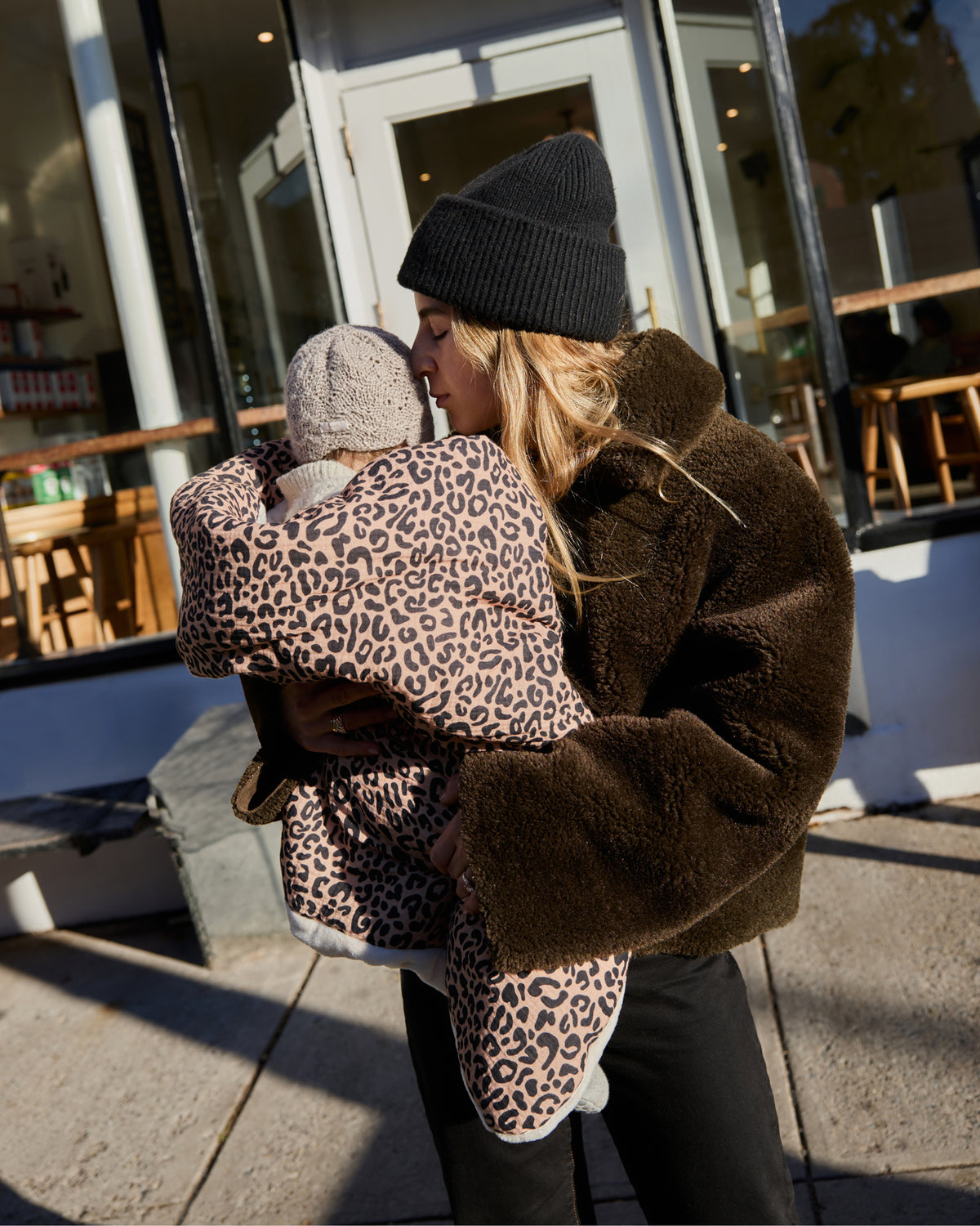 A woman outdoors in front of a cafe window, wearing a dark brown teddy coat and black beanie while holding a baby wrapped in Cozy Stroller Blanket in Leopard and a knitted hat.