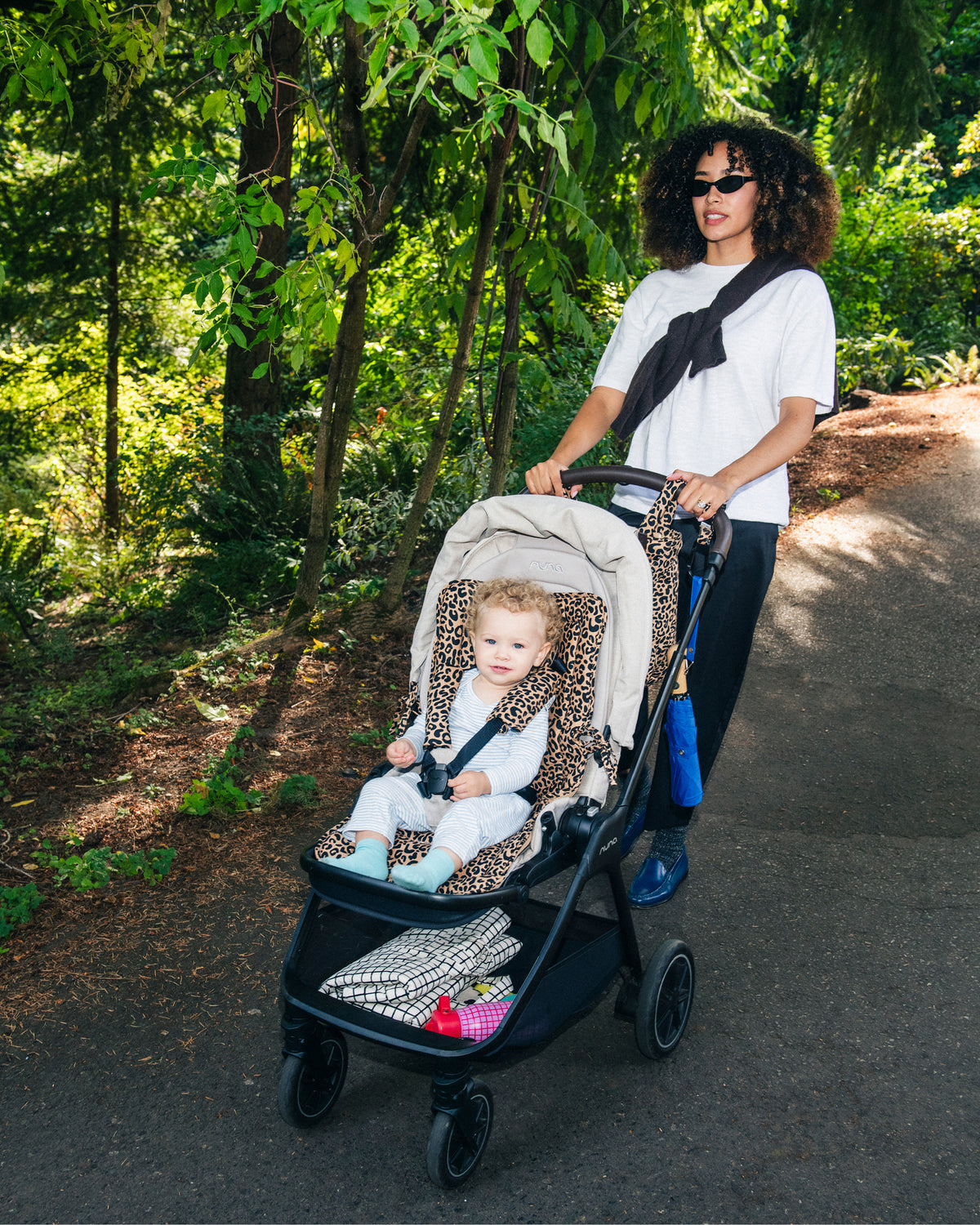 Model is walking a paved path through a park forest, pushing a baby in a grey stroller with Stroller Party™ Seat Liner & Strap Covers in Leopard with a Inside/Outside Play Mat in Black Grid and Cool Kid Water Bottle in Orchid Grid in the lower storage compartment. A Stroller Party™ Bucket Bag in Leopard and blue umbrella hang from the handlebar of the stroller.