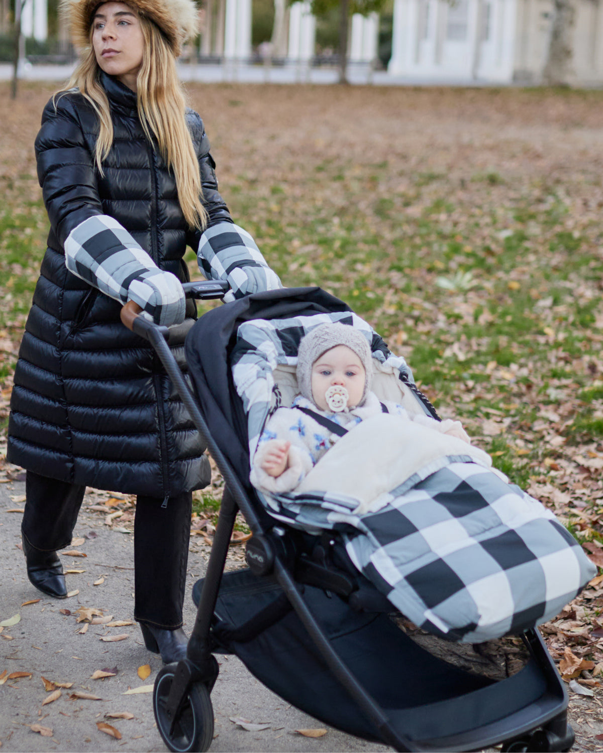A woman in a black puffer coat pushes a stroller outdoors on a leaf-covered path in a city park. The model pushes the stroller handlebar in the Cozy Stroller Mitts in Buffalo Plaid, and a baby is seated inside the Cozy Stroller Footmuff in Buffalo Plaid, while wearing a knit hat and pacifier.