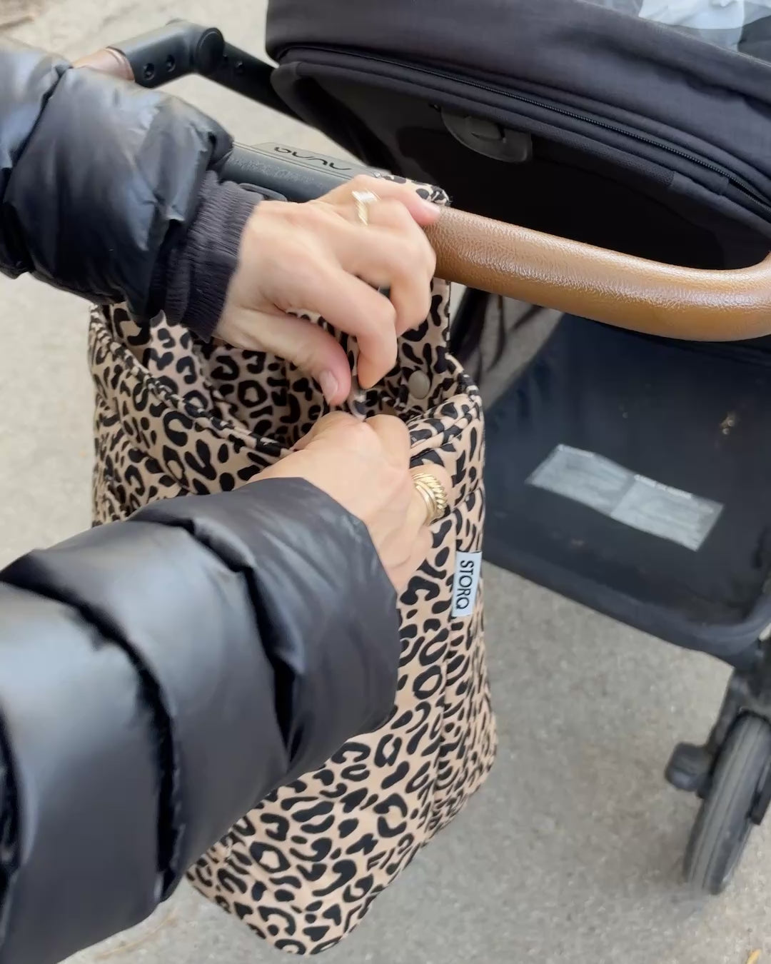 Movie of woman easily snapping straps back into Stroller Party™ Bucket Bag in Leopard, securing it to the handlebars of the black stroller while outdoors.