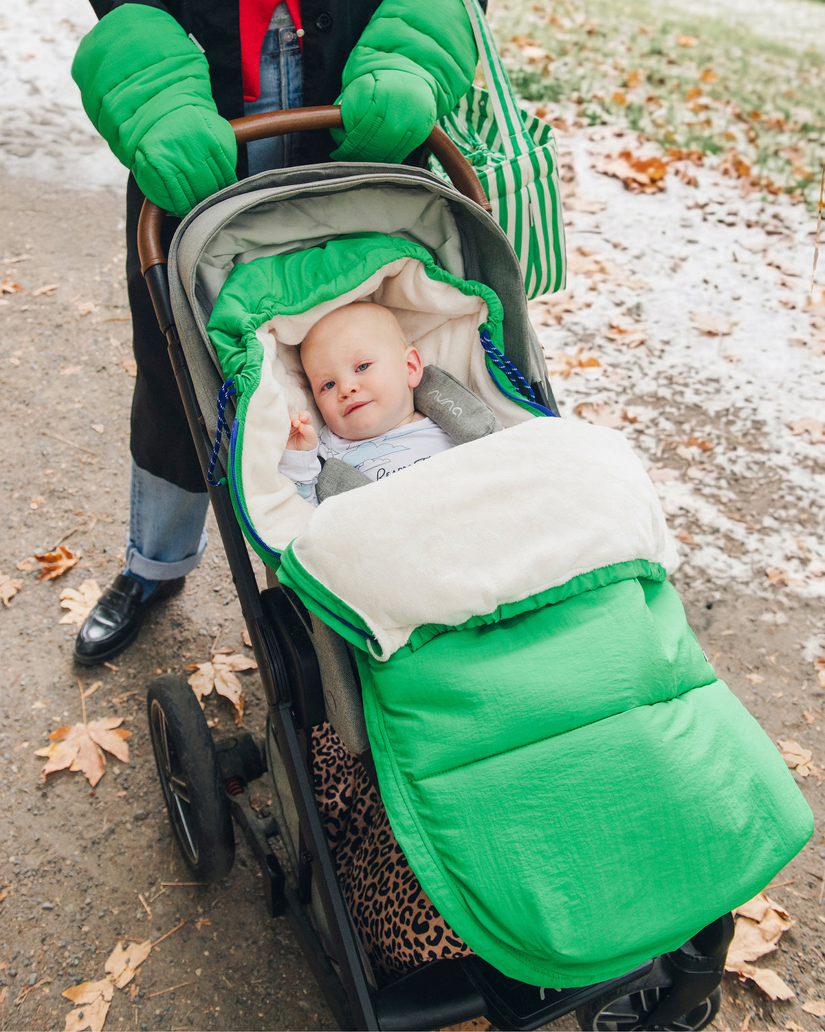 Mother is pushing baby in grey stroller through a park with snow on the ground. Baby is snuggled into the Cozy Stroller Footmuff in Green and the mother is wearing Cozy Stroller Mitts in Green, jeans, full black coat and Stroller Party™ Bucket Bag in Green Stripe.